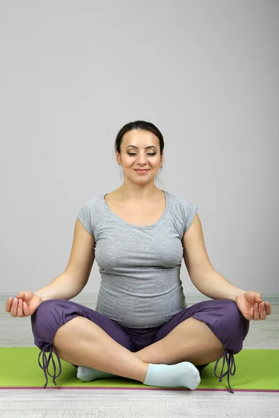 Mujer embarazada joven sentada y practicando yoga sobre estera sobre fondo de pared — Foto de Stock