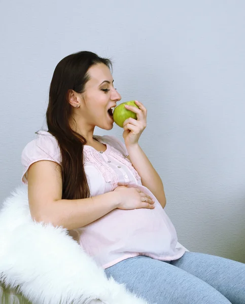 Mujer embarazada joven sentada en un sillón y comiendo manzana en el fondo de la pared — Foto de Stock