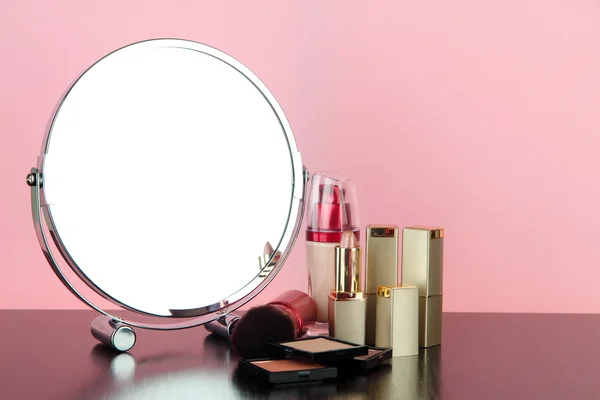 Round table mirror with cosmetics on table on pink background