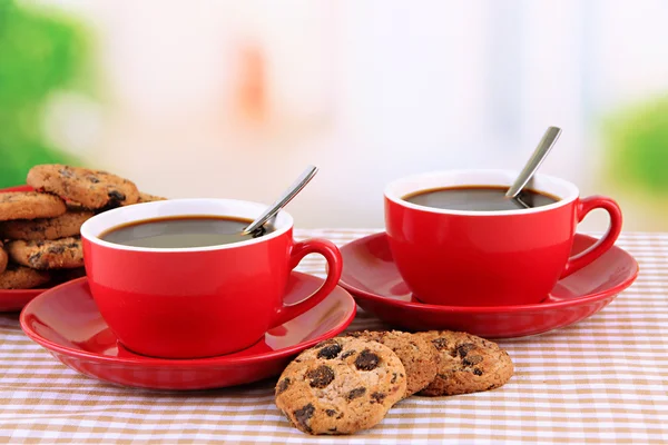 Copos vermelhos de café forte e biscoitos na toalha de mesa em fundo brilhante — Fotografia de Stock
