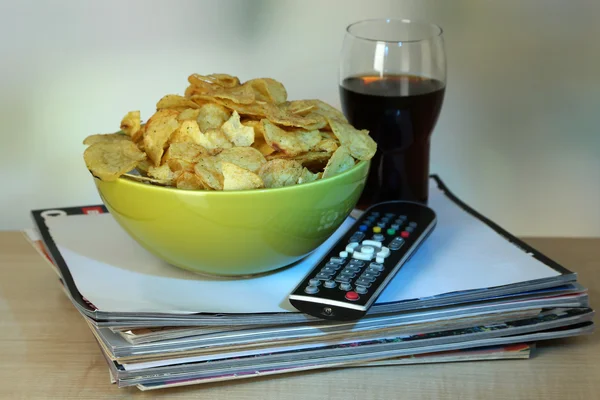 Chips in bowl, cola and TV remote on wooden table on room background — Stock Photo, Image