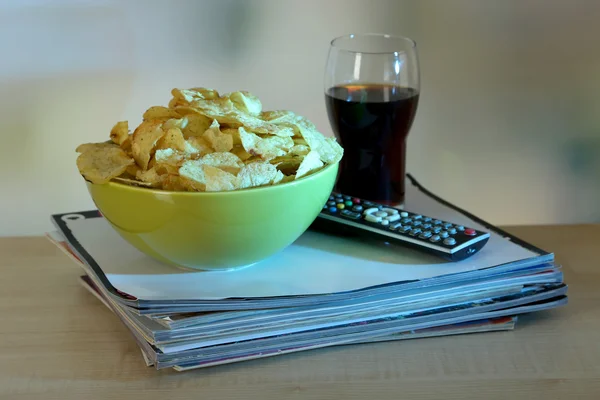 Chips en cuenco, cola y TV remoto en mesa de madera en el fondo de la habitación — Foto de Stock