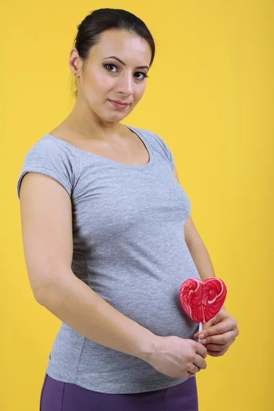 Young pregnant woman holding red lollipop on yellow background — Stock Photo, Image