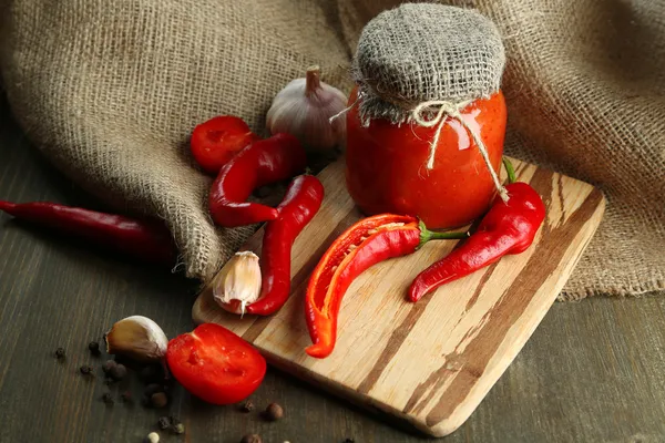 Composition with salsa sauce in glass jar,, red hot chili peppers and garlic, on sackcloth, on wooden background — Stock Photo, Image