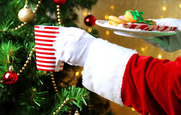 Santa holding mug and plate with cookies in his hand, on bright background — Stock Photo, Image