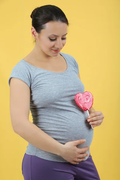 黄色の背景に赤の棒付きキャンデーを保持若い妊娠中の女性 — ストック写真