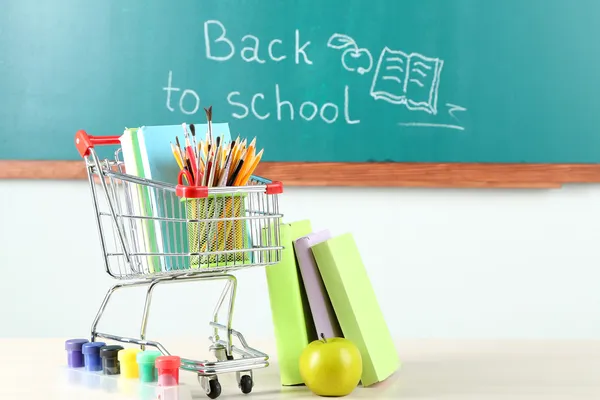 School supplies in supermarket cart on blackboard background — Stock Photo, Image