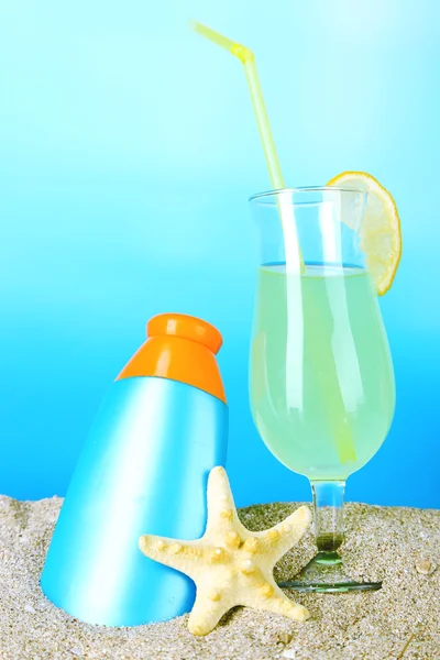 Beach cocktail and sunscreen in sand on blue background — Stock Photo, Image