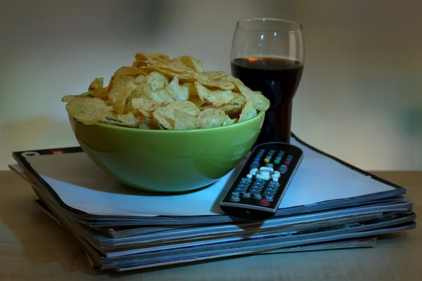 Chips in bowl, cola and TV remote on wooden table on room background — Stock Photo, Image