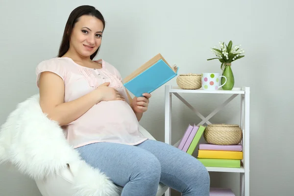 Nuori regnant nainen istuu nojatuoli ja lukeminen kirja seinällä tausta — kuvapankkivalokuva