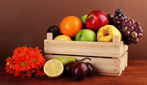 Verschillende vruchten in krat en bloemen op tafel op bruine achtergrond — Stockfoto
