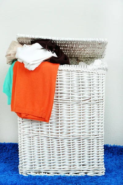 Cesta de lavandaria completa no tapete de cor no fundo da parede cinza — Fotografia de Stock