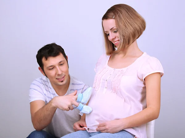 Young pregnant woman with her husband holding baby shoes on gray background — Stock Photo, Image