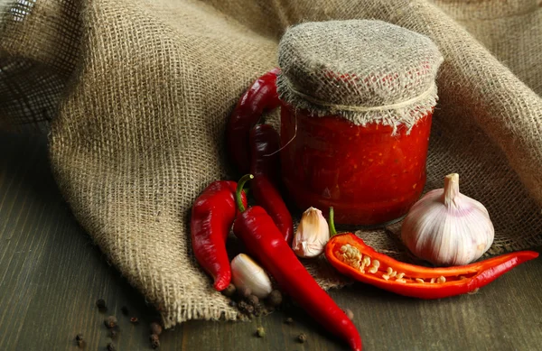 Composizione con salsa in vaso di vetro, peperoncino rosso e aglio, su sacco, su fondo di legno — Foto Stock