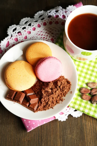 Cocoa in cup with sweets and cocoa powder on plate on wooden table — Stock Photo, Image