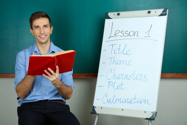 Young teacher sitting near chalkboard in school classroom — Stock Photo, Image