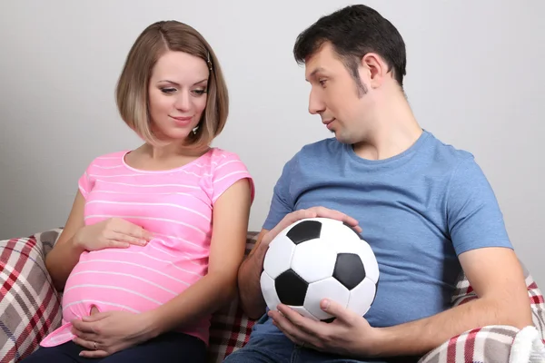 Mladá těhotná žena a její manžel drží míč na gauči na šedém pozadí — Stock fotografie