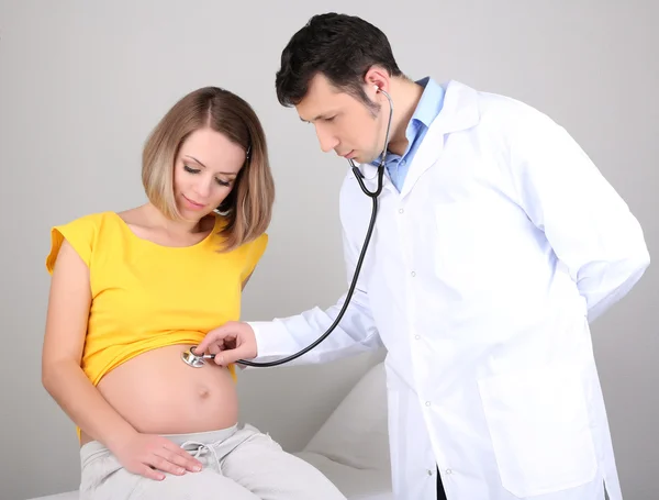 Ung gravid kvinna som sitter på sjukhussäng med läkare på grå bakgrund — Stockfoto