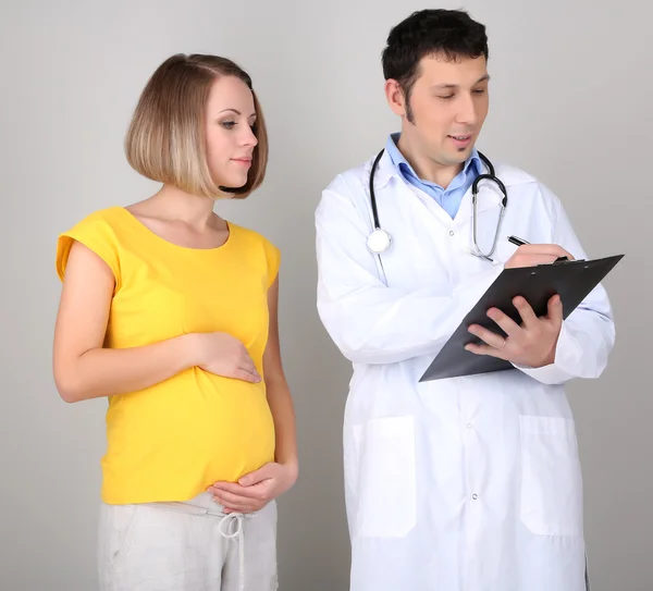 Jovem grávida com médico em fundo cinza — Fotografia de Stock