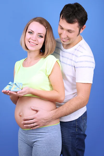 Jeune femme enceinte tenant des chaussures de bébé avec son mari sur fond bleu — Photo