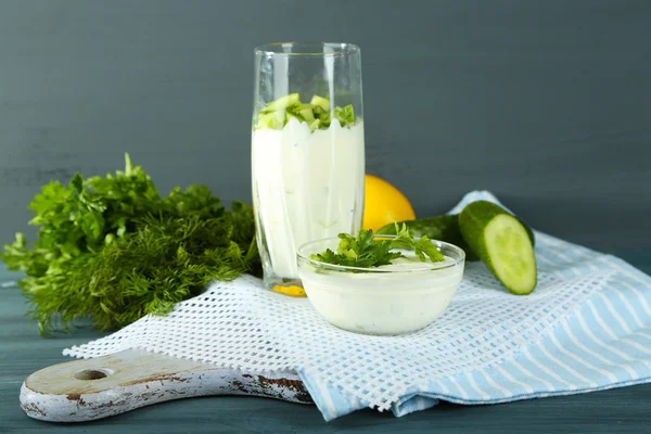 Cucumber yogurt in glass and bowl, on color napkin, on wooden table, on gray background — Stock Photo, Image