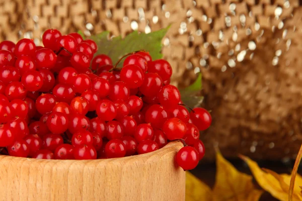 Красные ягоды вибурнума в деревянной чаше с желтыми листьями на столе на коричневом фоне — стоковое фото