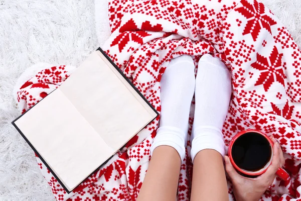 Состав с теплым пледом, книга, чашка горячего напитка и женские ноги, на фоне цветного ковра — стоковое фото