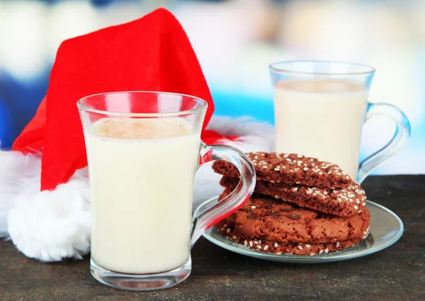 Cups of eggnog with cookies and Santa hat on table on bright background — Stock Photo, Image