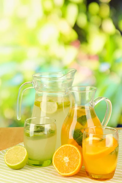 Limonata di arancia e limone in brocche e bicchieri su tavolo in legno su sfondo naturale — Foto Stock