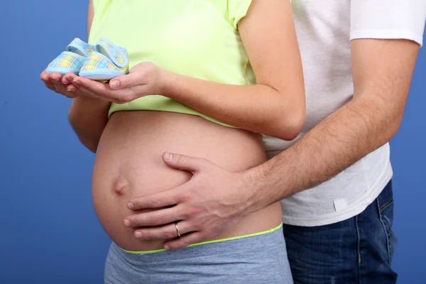 青色の背景に彼女の夫と赤ちゃんの靴を保持している妊娠中の女性 — ストック写真
