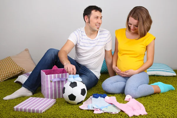 Young pregnant woman with her husband folding baby wear and ball on floor at home — Zdjęcie stockowe