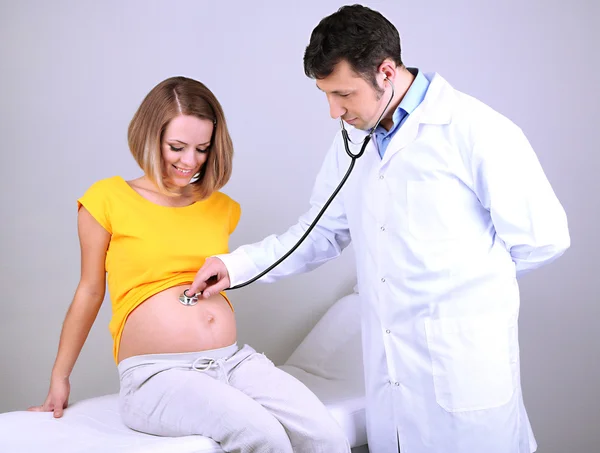 Young pregnant woman sitting on hospital bed with doctor on gray background — Stock Photo, Image