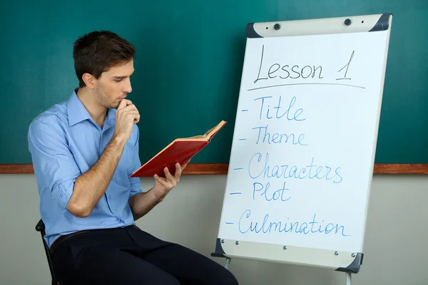 Young teacher sitting near chalkboard in school classroom — Stock Photo, Image