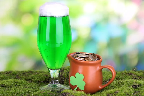 Glass of green beer and pitcher with coins on grass on natural background — Stock Photo, Image