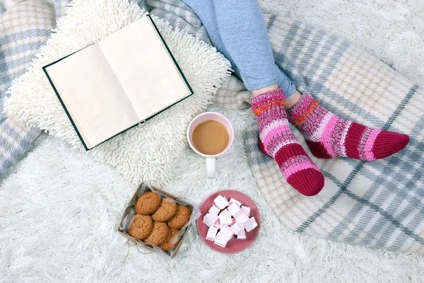 Composizione con plaid caldo, libro, tazza di bevanda calda e gambe femminili, su sfondo tappeto di colore — Foto Stock