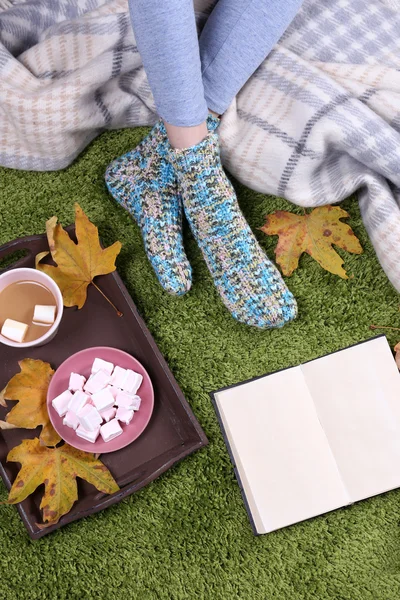 Složení s teplý pléd, kniha, šálek horké nápoje a ženské nohy, na koberec barvy pozadí — Stock fotografie