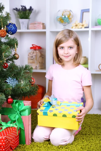 Little girl holding present box near Christmas tree in room Stock Picture