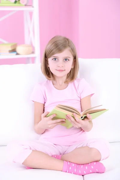 Lilla flickan läsa bok sittande på soffan i rummet — Stockfoto