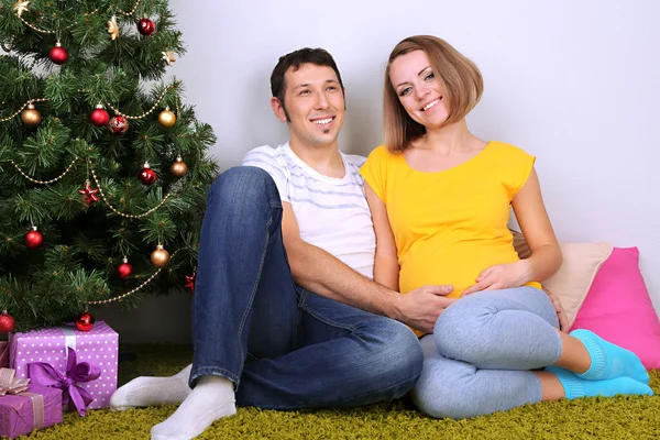 年轻孕妇和她的丈夫坐在圣诞树在家附近的地板上 — 图库照片