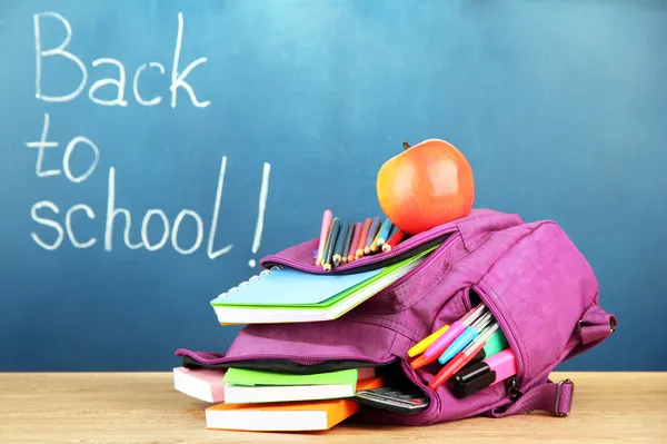 Purple backpack with school supplies on wooden table on green desk background — Stock Photo, Image