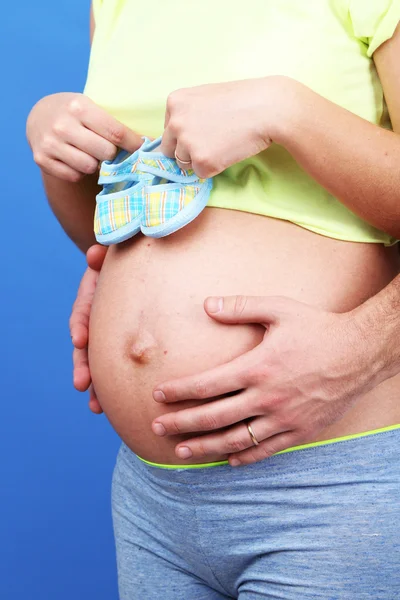 Pregnant woman holding baby shoes with her husband on blue background — Stock Photo, Image