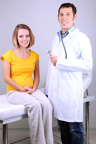 Jonge zwangere vrouw zittend op ziekenhuisbed met arts op grijze achtergrond — Stockfoto