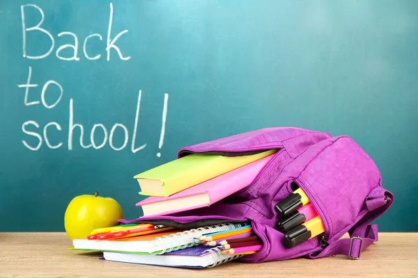 Lila ryggsäck med skolan levererar på träbord på gröna skrivbord bakgrund — Stockfoto
