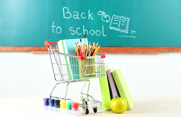 School supplies in supermarket cart on blackboard background — Stock Photo, Image