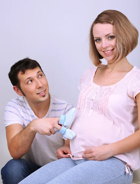 Young pregnant woman with her husband holding baby shoes on gray background — Stock Photo, Image