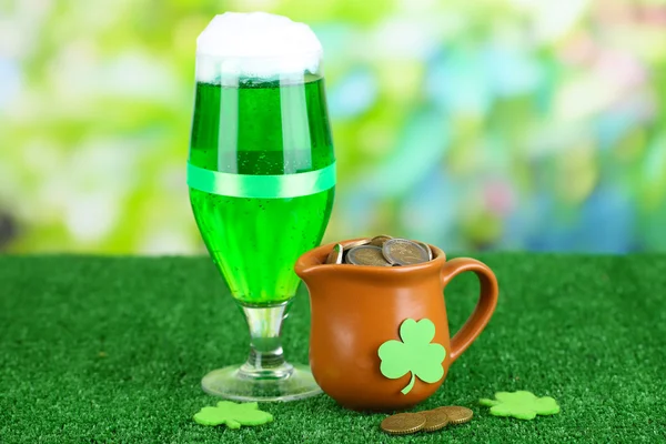 Glass of green beer and pitcher with coins on grass on natural background — Stock Photo, Image