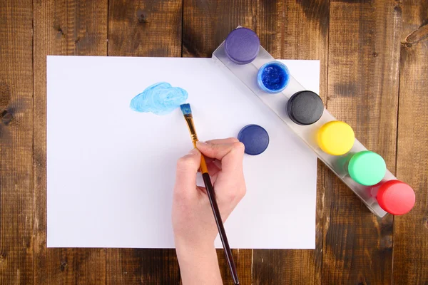 Hand holding brush with paints and paper on wooden background — Stock Photo, Image