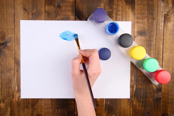 Hand som håller penseln med färger och papper på trä bakgrund — Stockfoto