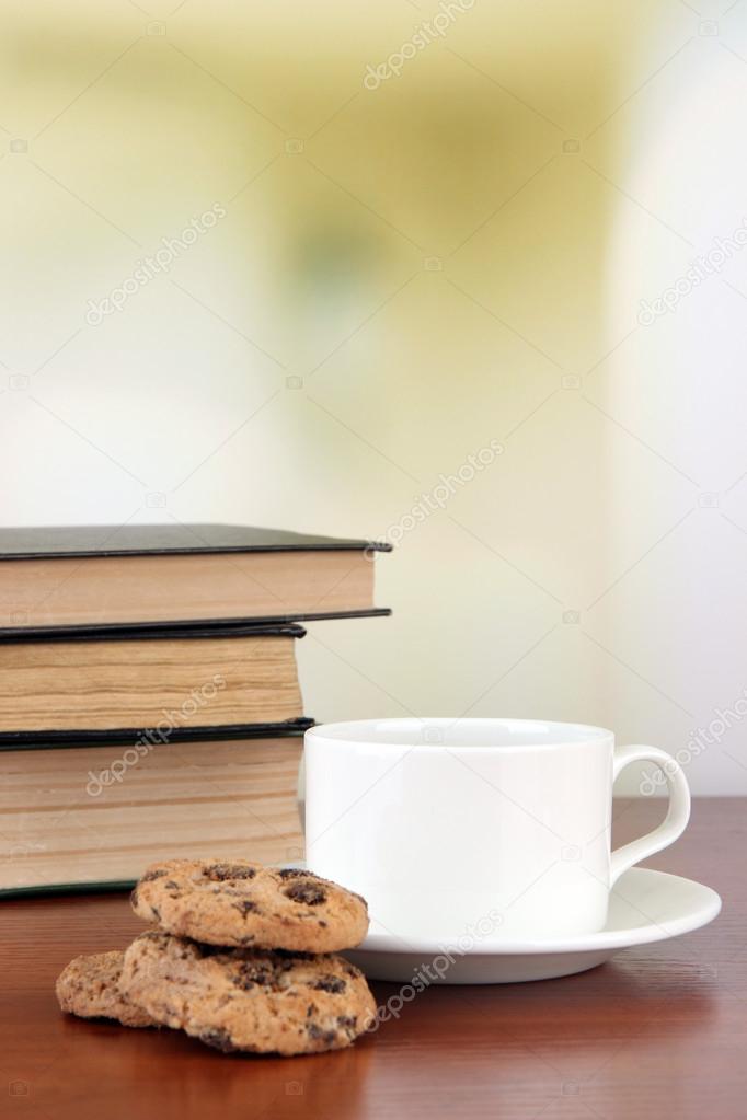 Cup of coffee with cookies and books on wooden table on bright background