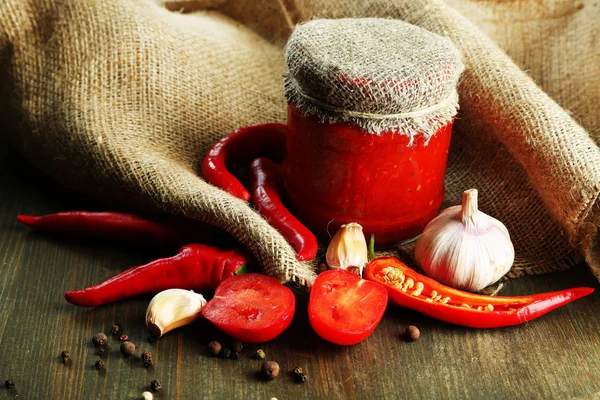 Composition avec sauce salsa dans un bocal en verre, piments rouges et ail, sur un sac, sur un fond en bois — Photo